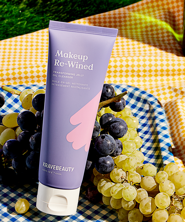 A purple tube of KraveBeauty Makeup Re-Wined cleanser resting on a bunch of grapes on a picnic blanket.