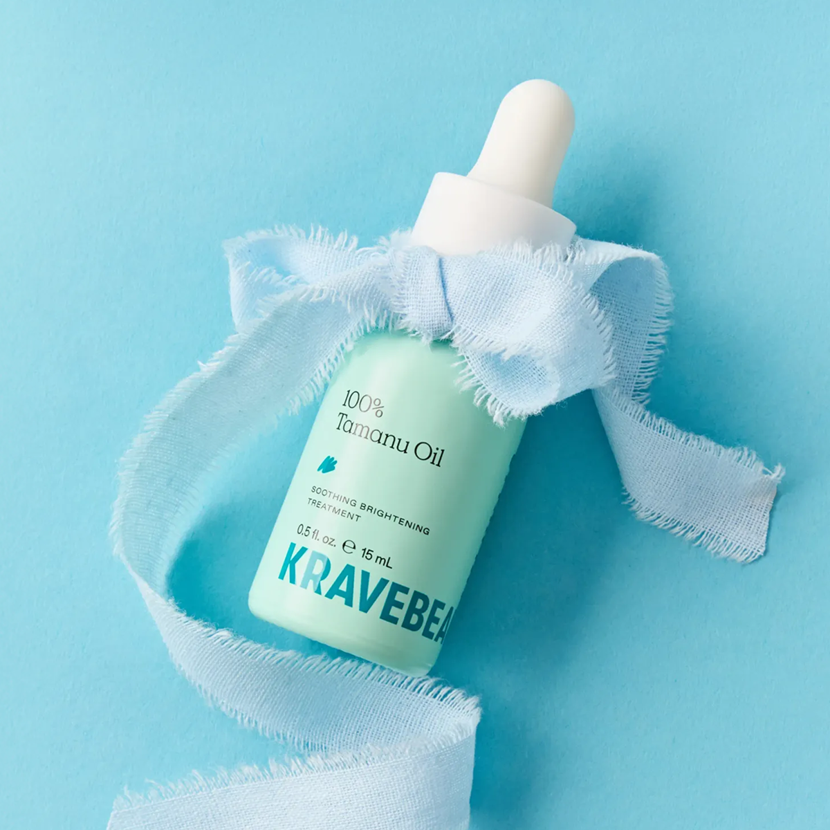 A bottle of KraveBeauty Tamanu Oil with a light blue frayed ribbon tied around it on a blue background.
