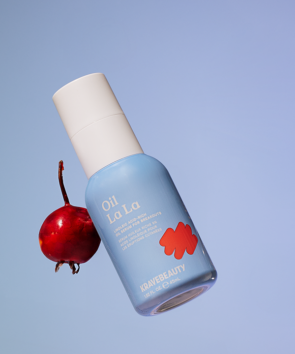 A light blue bottle of KraveBeauty Oil La La serum floats next to a red berry against a blue background.