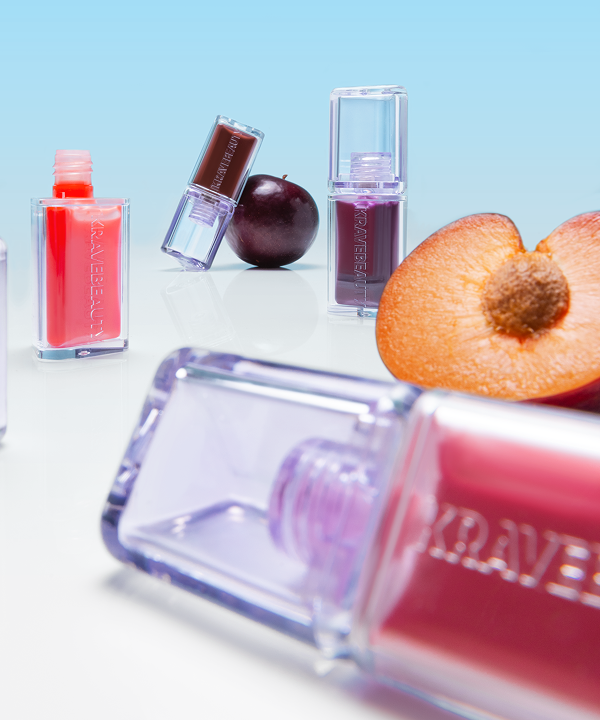 Several bottles of KraveBeauty lip tint are arranged on a reflective surface with whole and sliced plums.