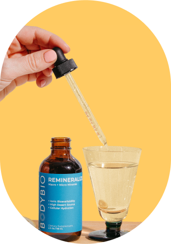 A hand using a dropper with a glass of liquid and a bottle labeled 'BODYBIO REMINERALIZE.'