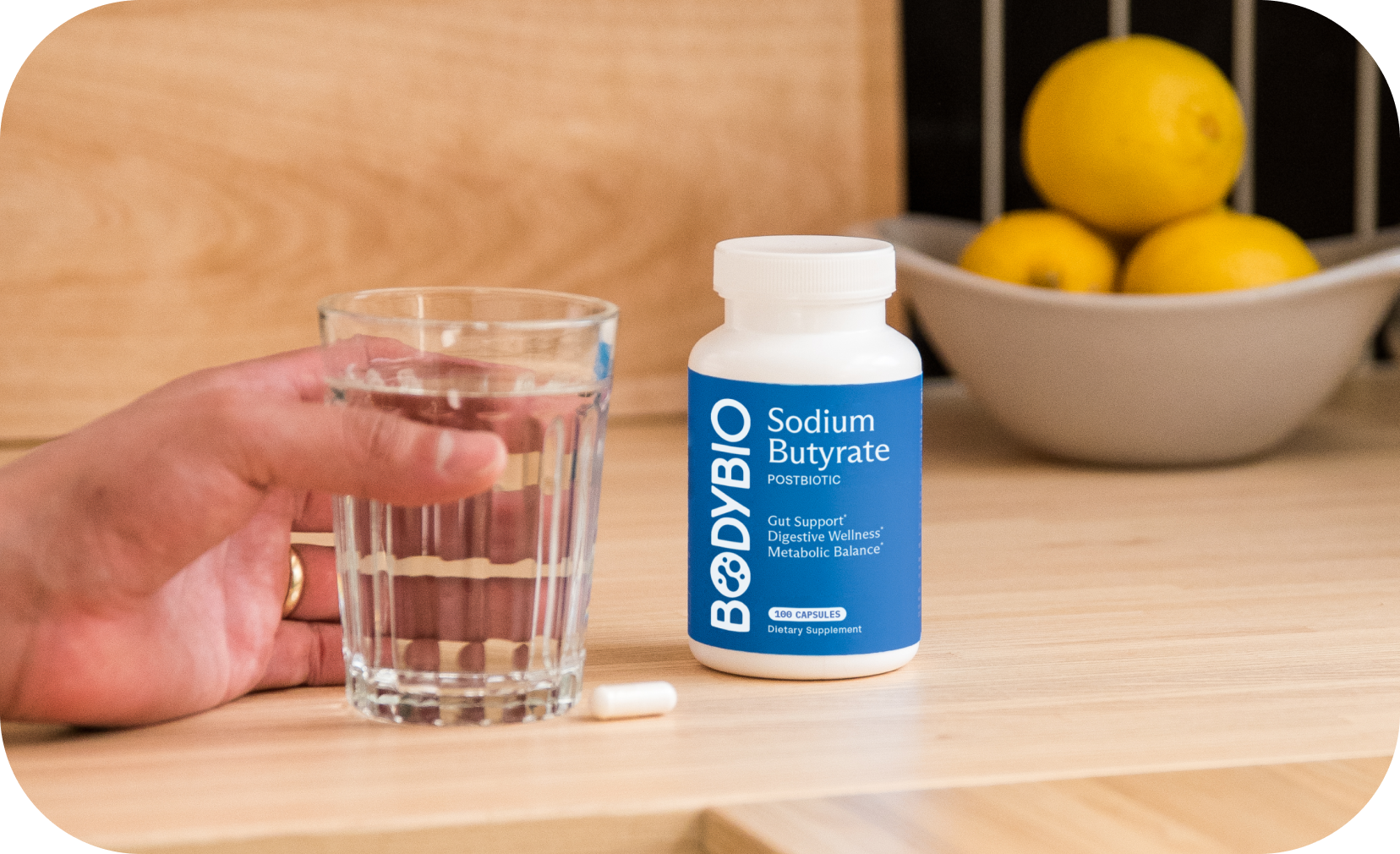 Hand holding a glass of water next to a bottle of sodium butyrate capsules.