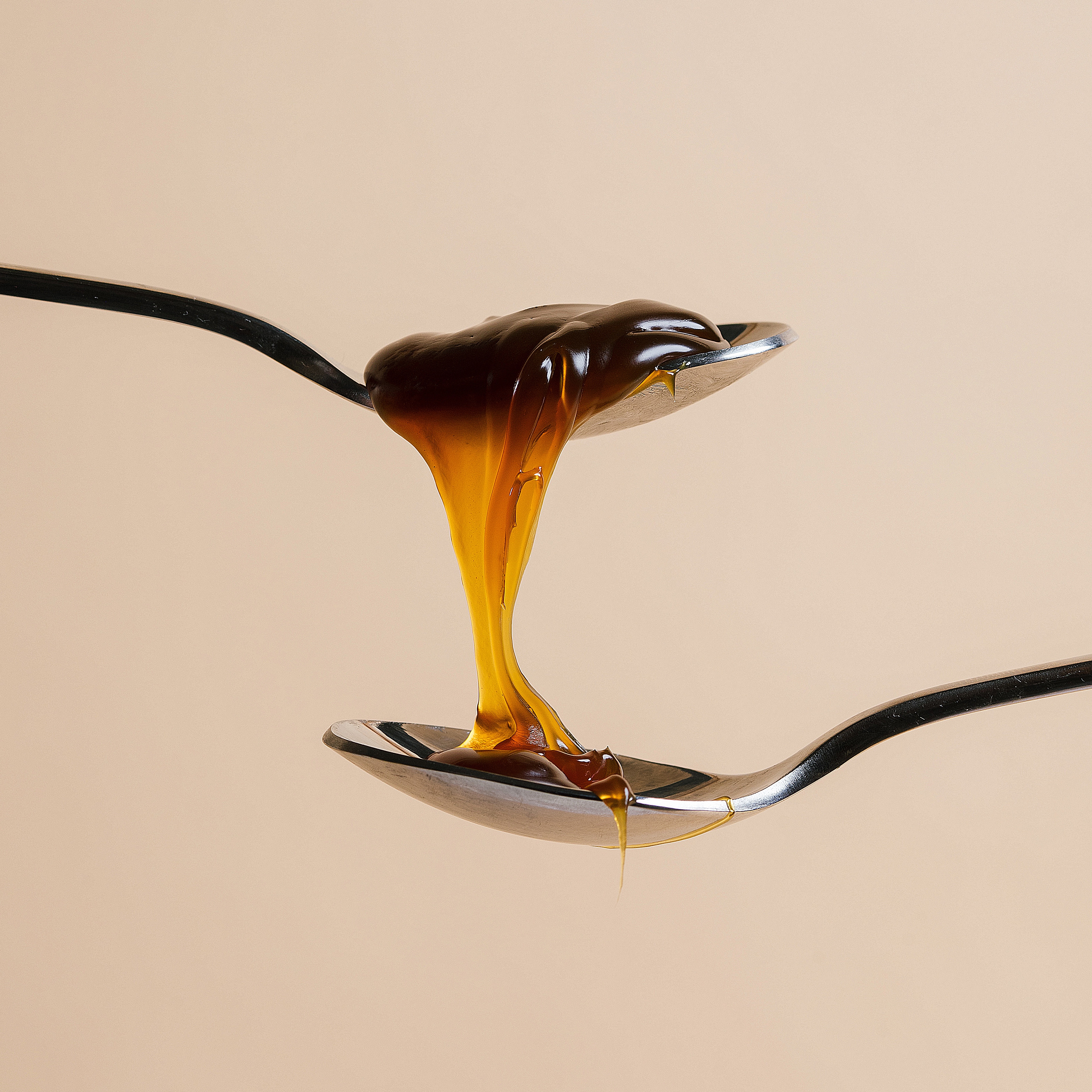 Thick, dark caramel sauce dripping from an upper silver spoon to a lower one against a beige background.
