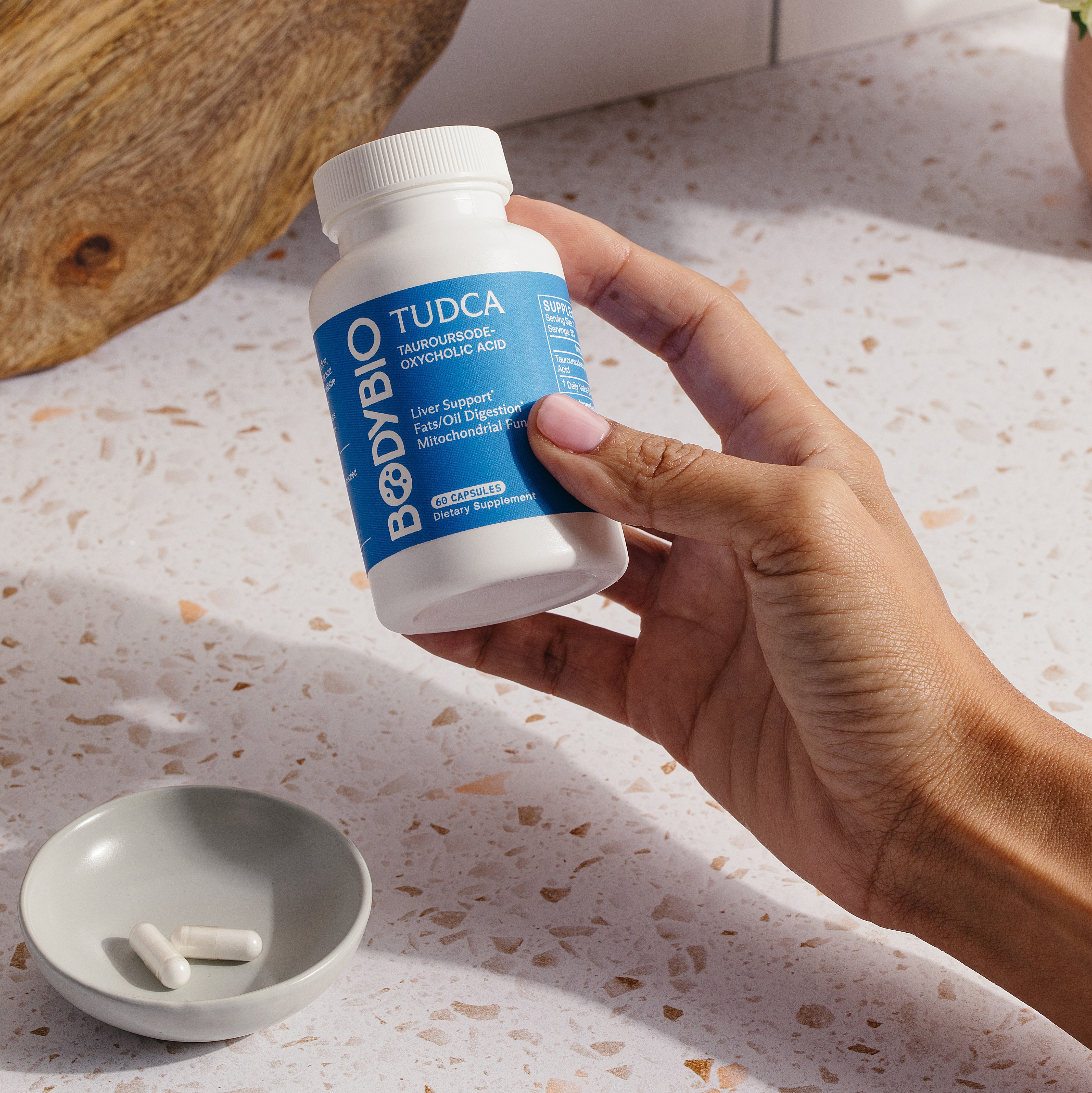 A hand holding a bottle of BodyBio TUDCA supplements, with two capsules resting in a small dish.