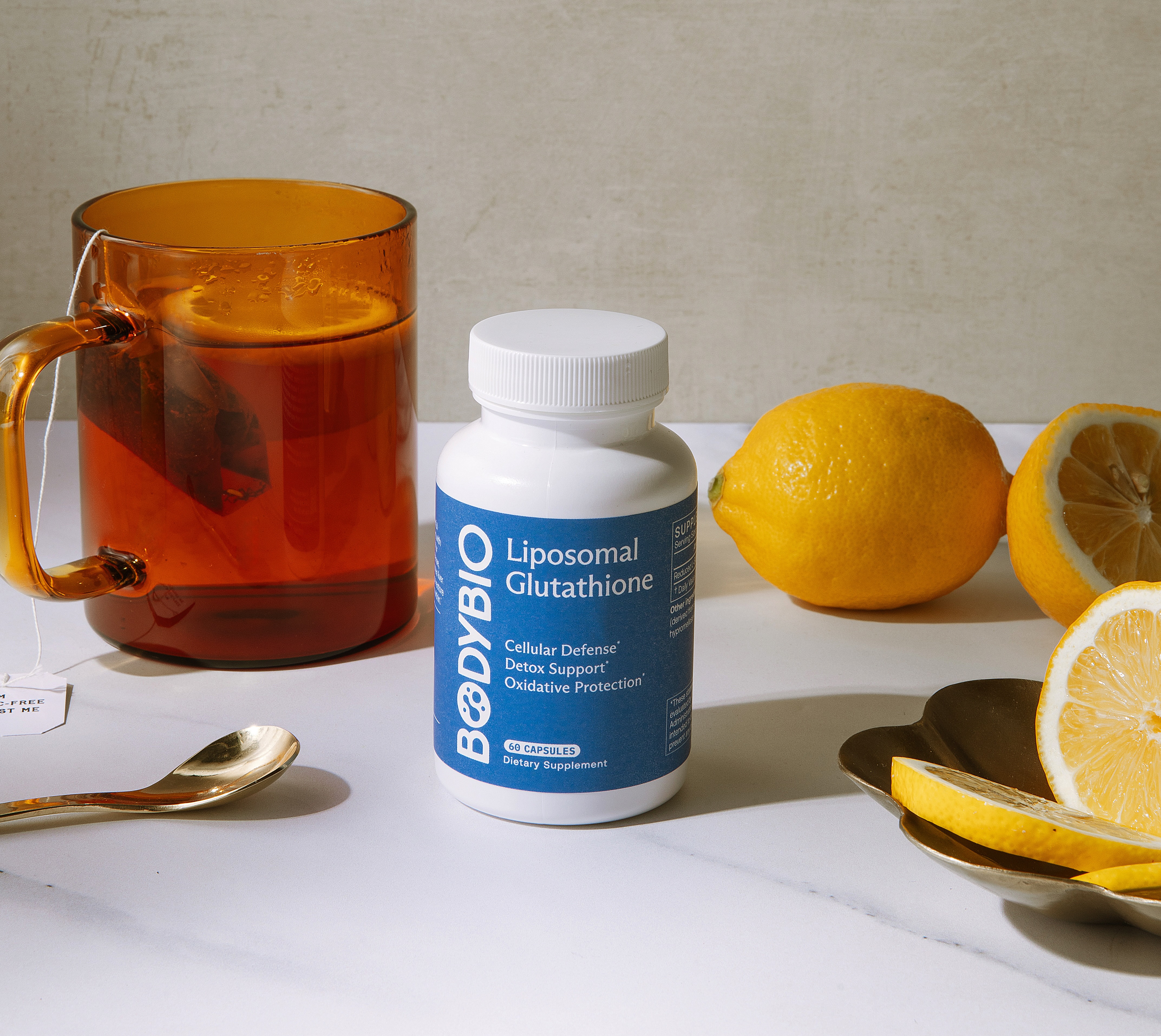 A bottle of BodyBio supplements sits on a marble surface next to a cup of tea and lemons.