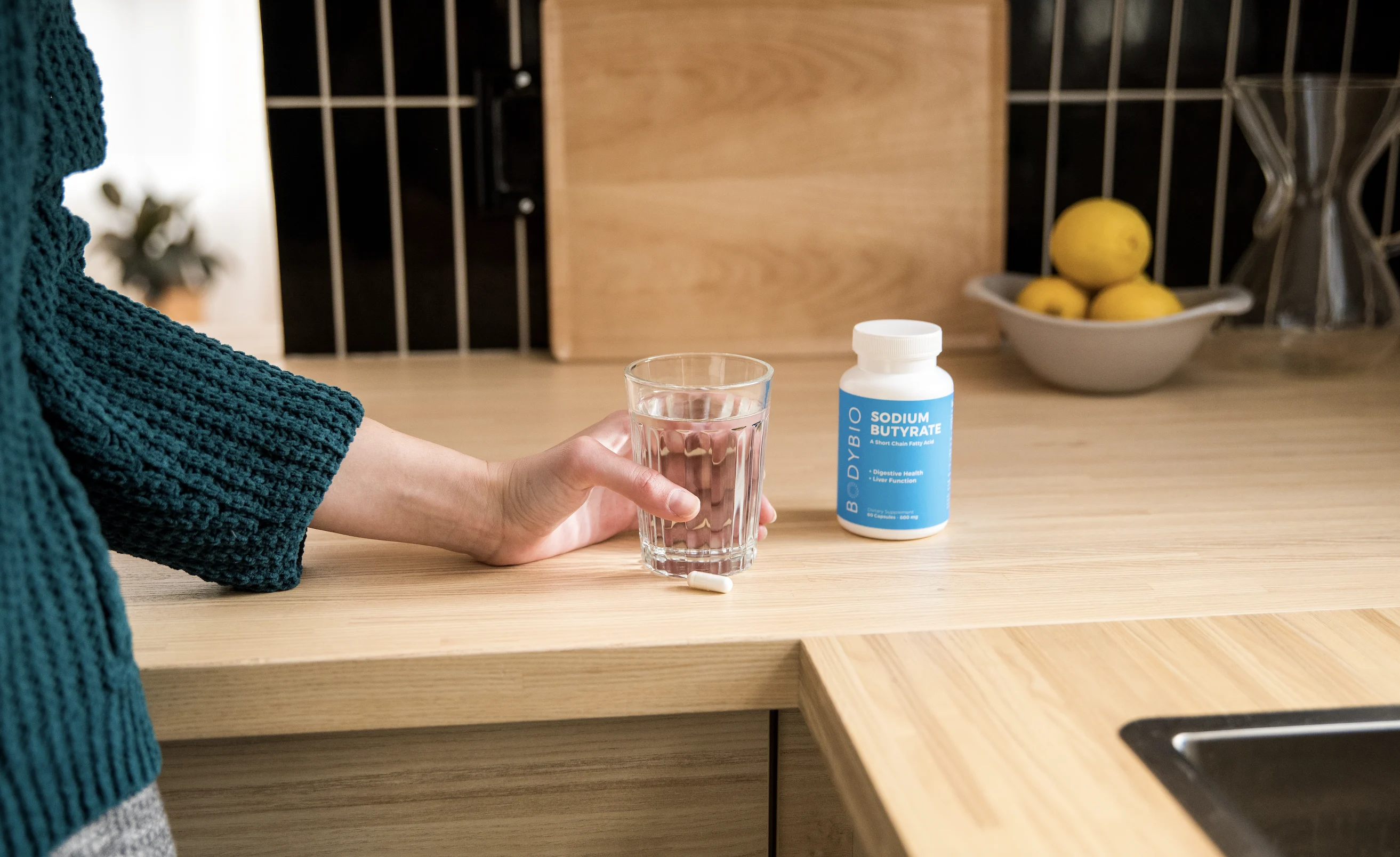 Hand holding a glass of water next to a pill and supplement bottle on a kitchen counter.