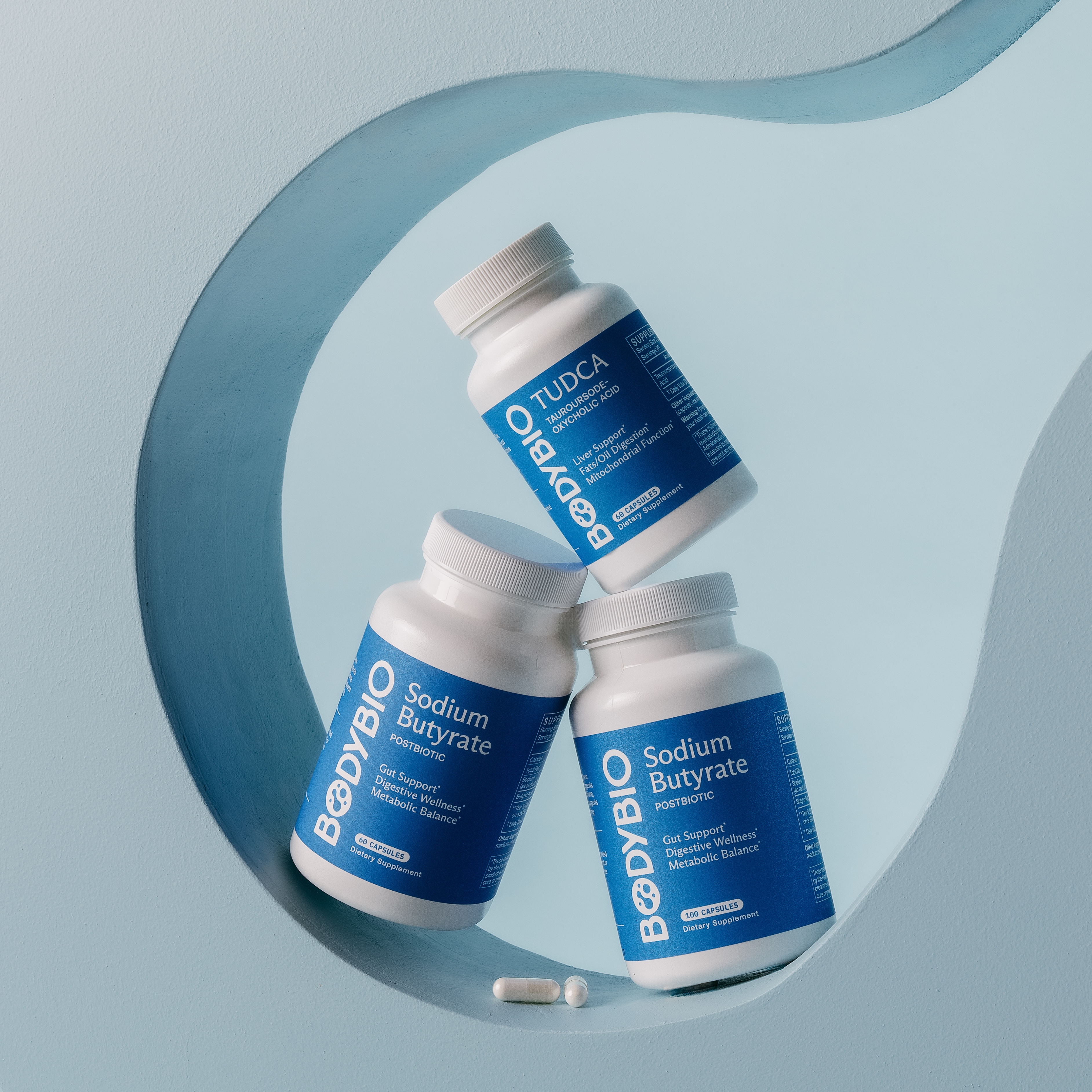 Three bottles of Bodybio TUDCA and Sodium Butyrate supplements on a stylized blue background with two capsules.