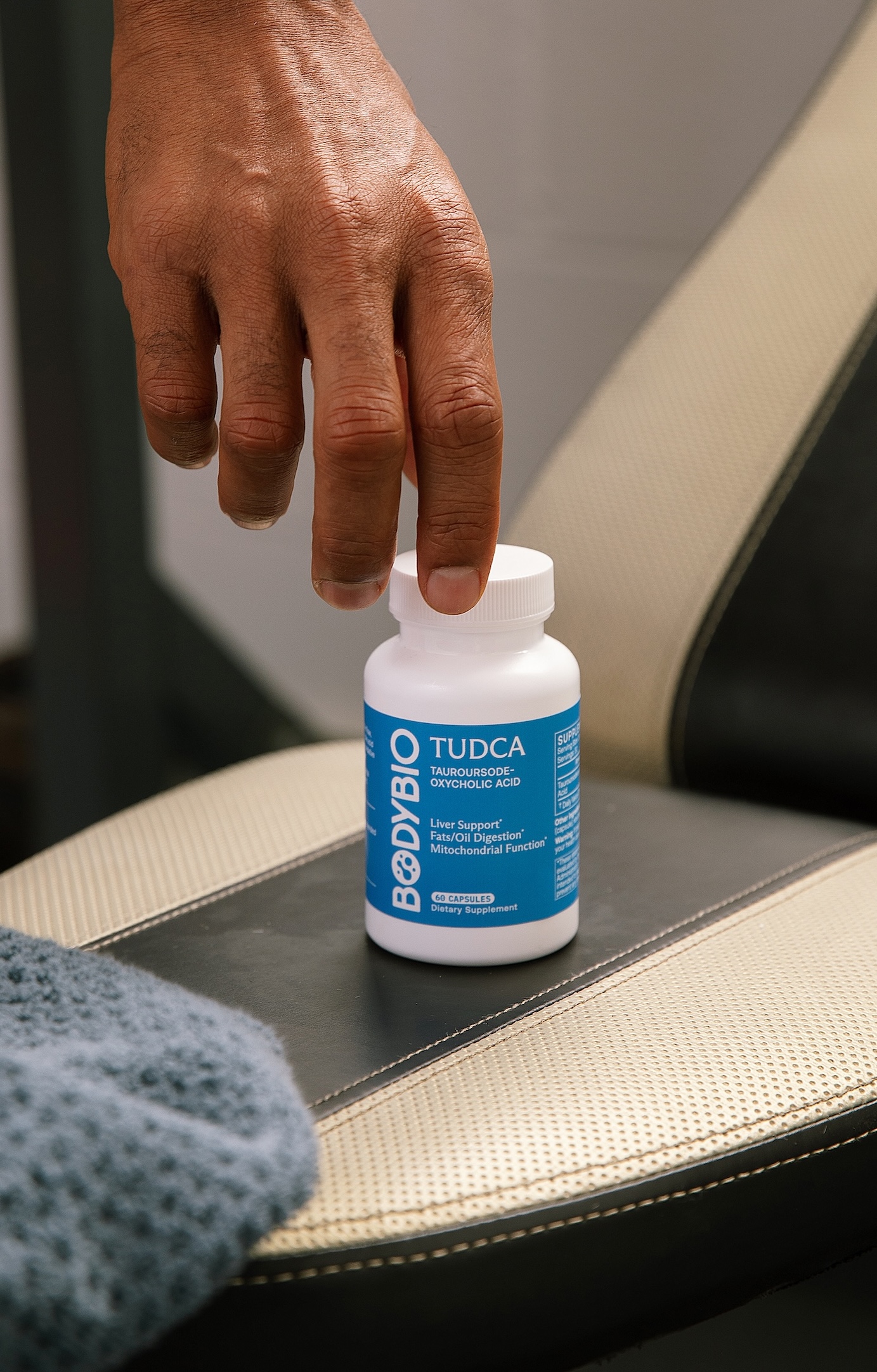 A person's hand reaches for a white bottle of BodyBio TUDCA supplements sitting on a chair.