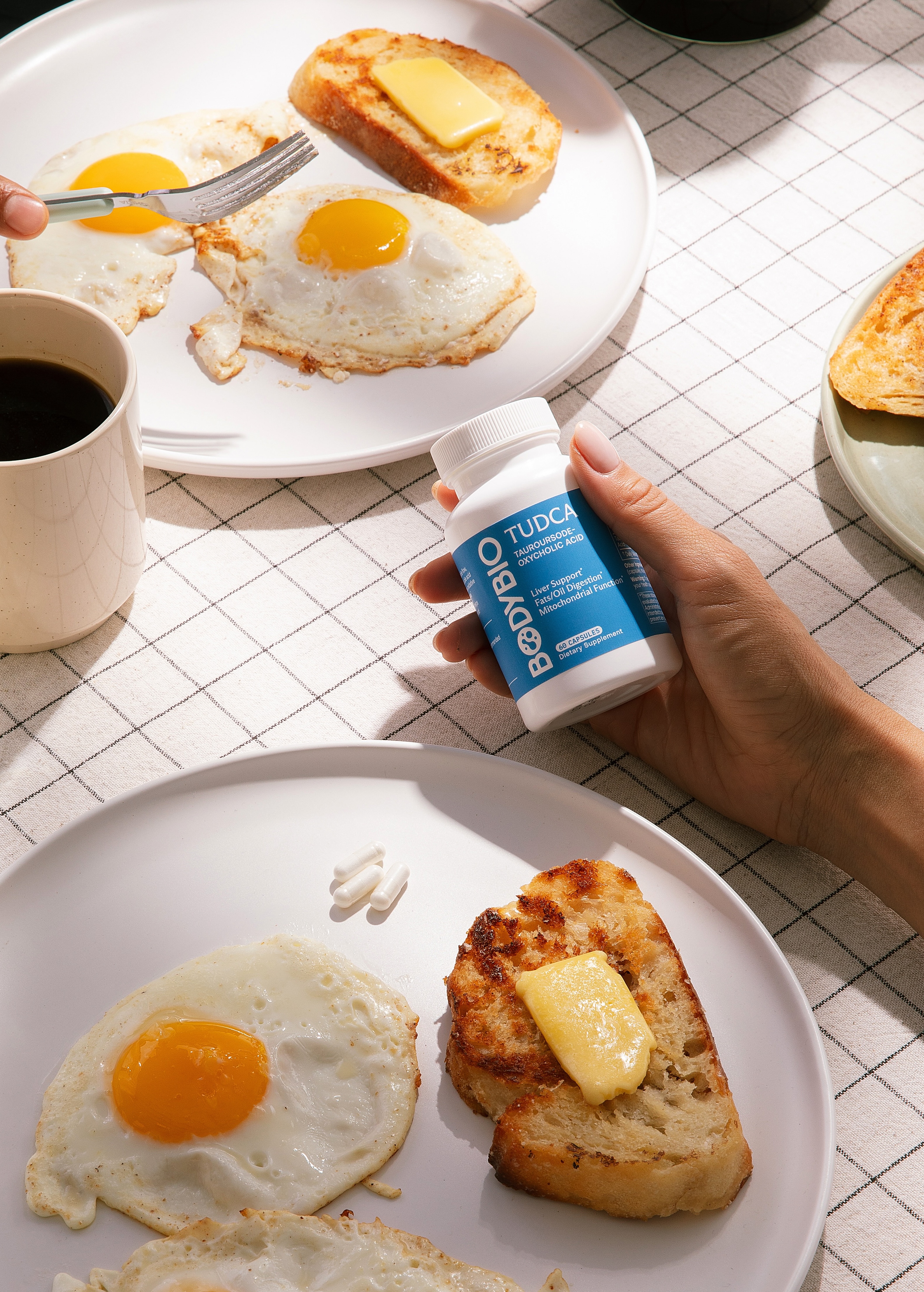 A person holds a bottle of BodyBio TUDCA supplements over a breakfast of fried eggs and toast.