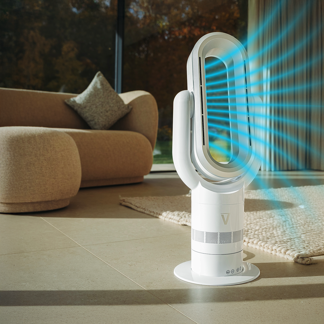A modern, white bladeless fan stands on a tiled floor in a sunlit living room, blowing air.