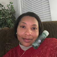 Person sitting on a couch holding a green bottle, with a plant and window blinds in the background.