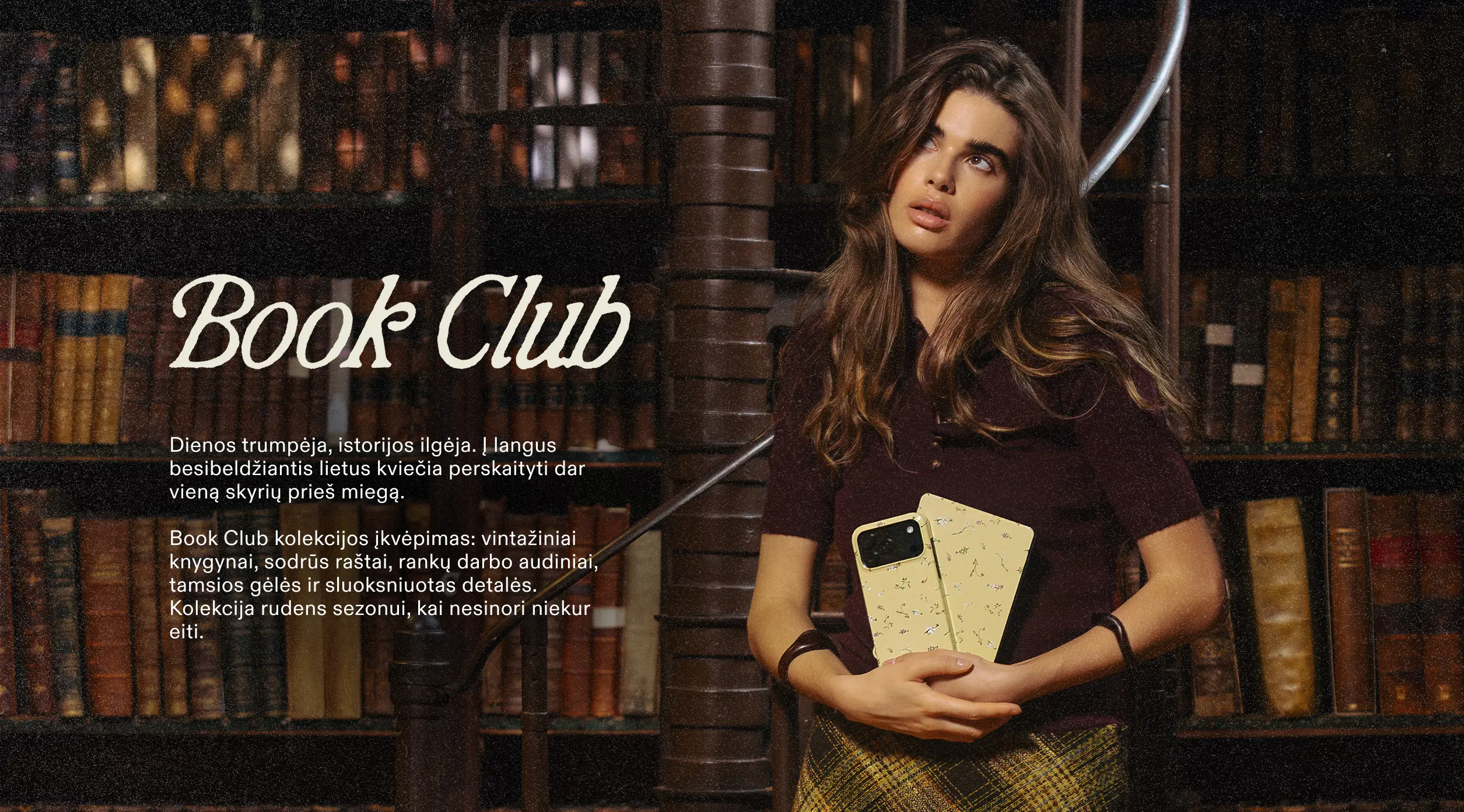 A woman with long brown hair stands in a library, holding a yellow patterned phone case and looking up.