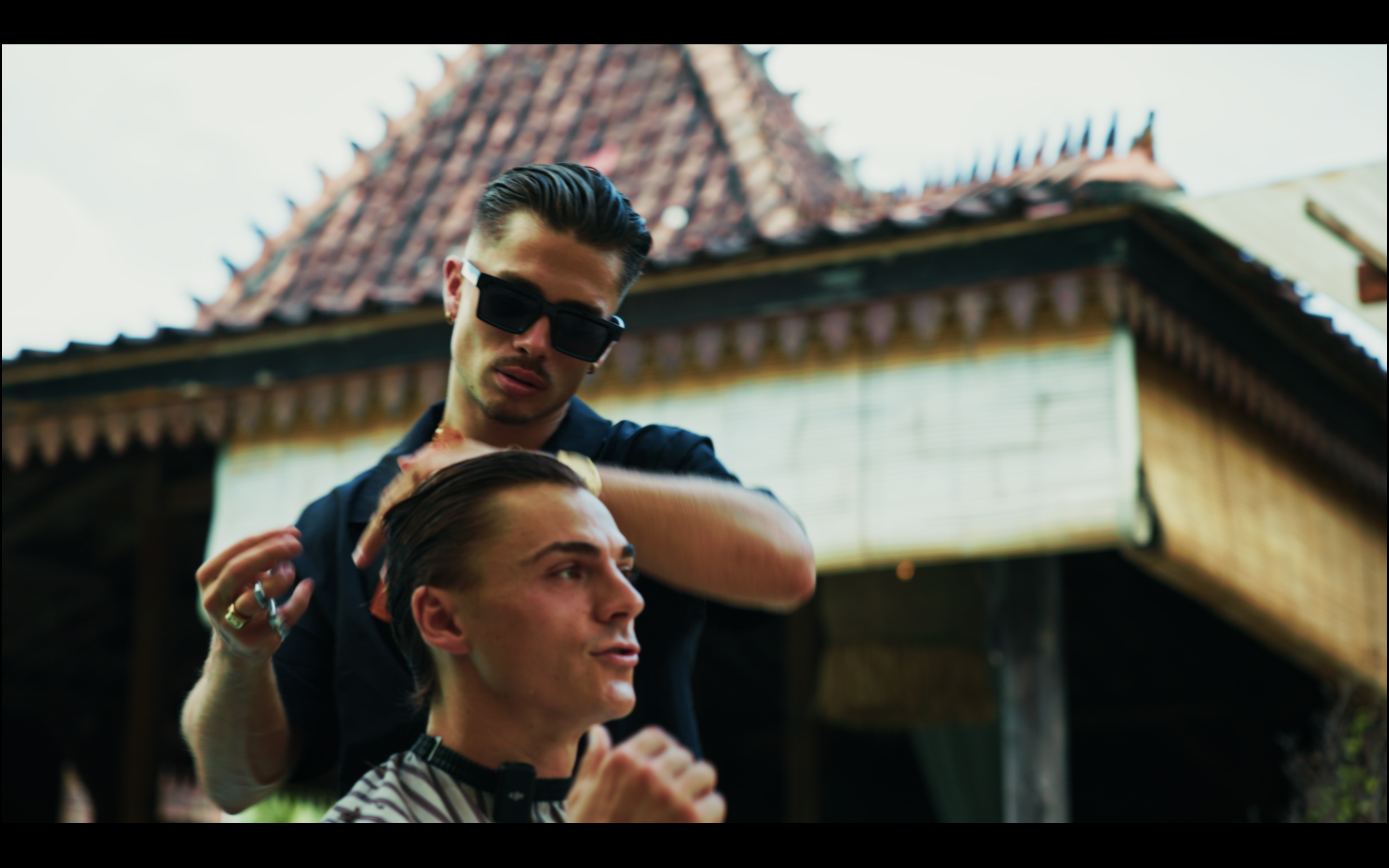 A barber styling a man's hair outdoors, with a traditional building in the background.