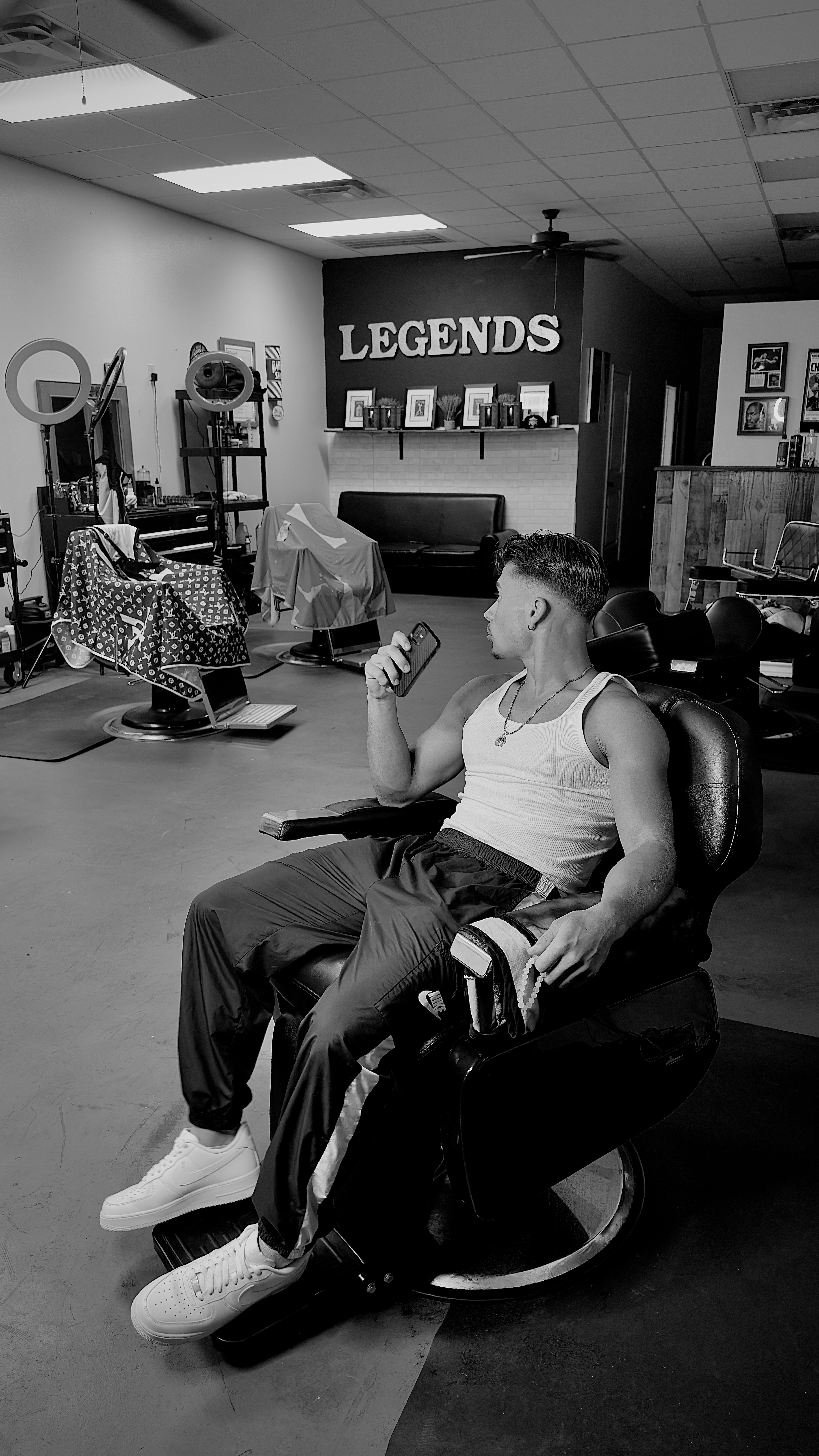 Man sitting in a barbershop chair, holding a phone, with 'Legends' sign in background.