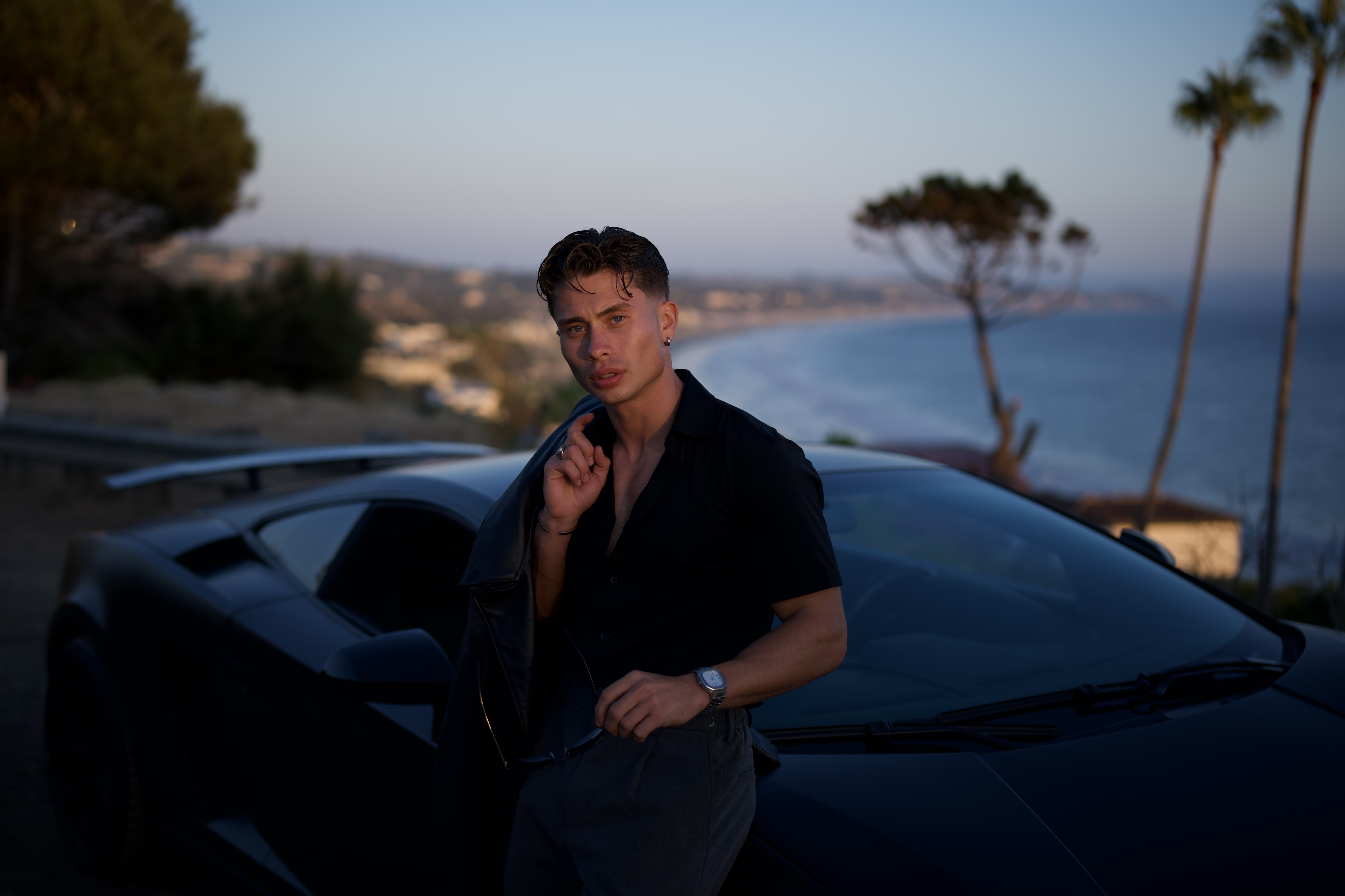 Person standing by a black sports car with ocean view in the background.