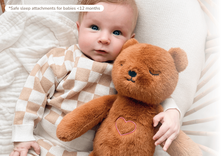 An overhead view of a baby with blue eyes hugging a brown teddy bear with a heart on its chest.