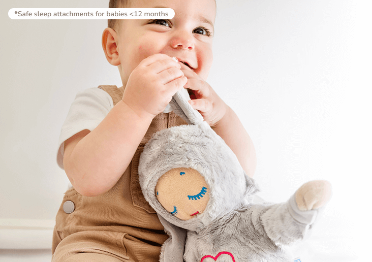 A smiling baby in brown overalls holds a grey plush toy to its mouth.