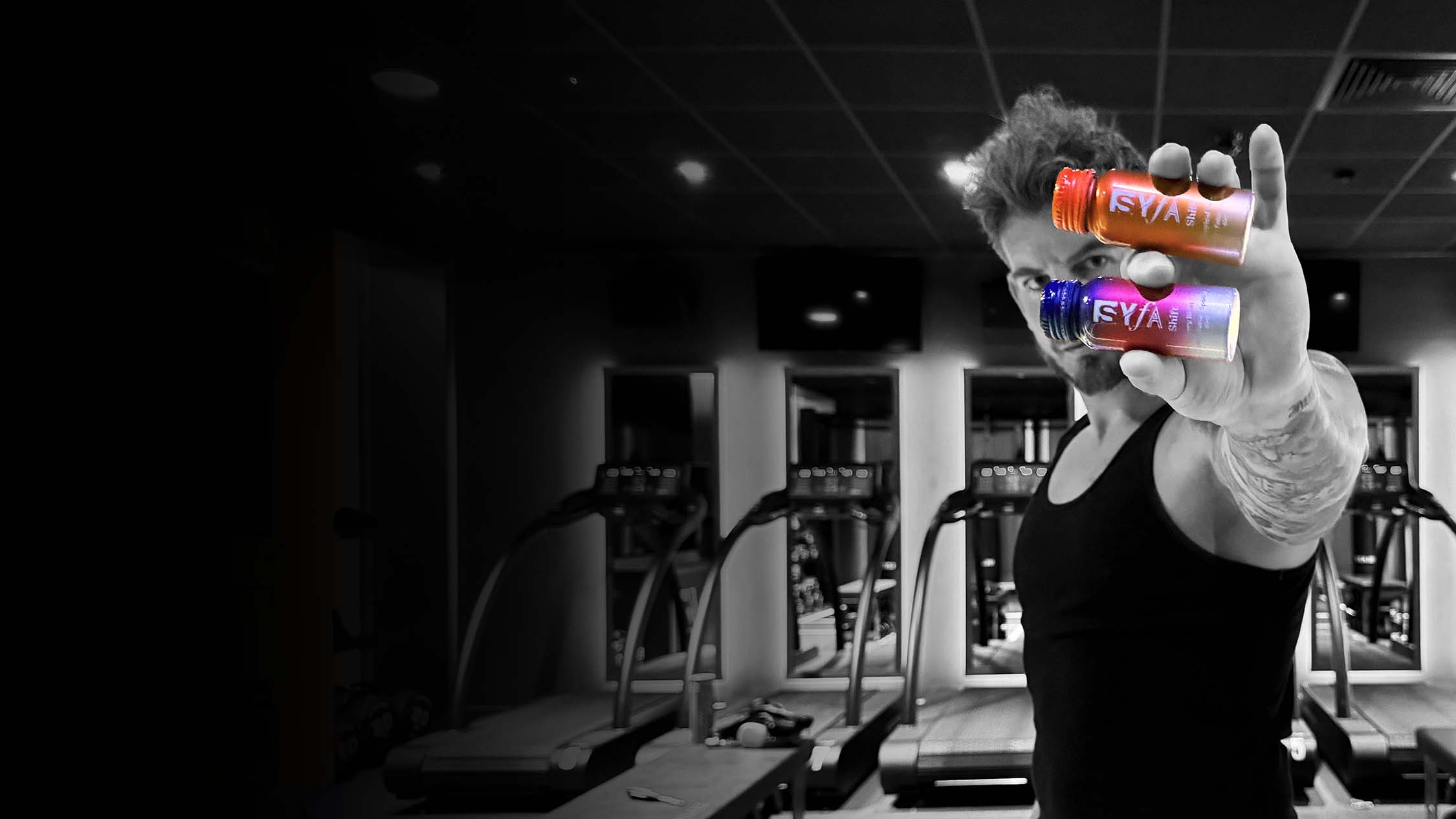 Man in gym holding two colorful bottles, treadmills in background.