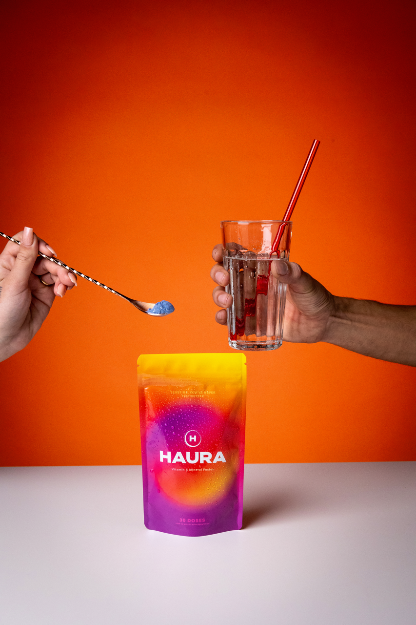 Hands holding a glass of water and a spoon with powder, next to a colorful Haura package.