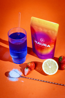 Colorful drink mix with glass, strawberries, lemon, and spoon on orange background.
