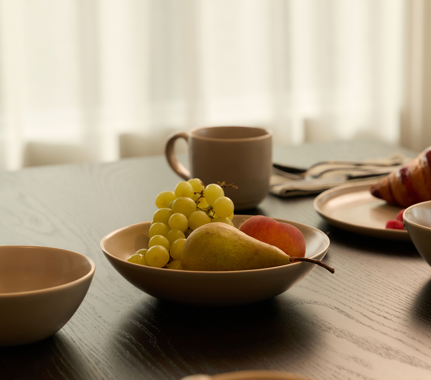 Ceramic bowl with fruit on a dining table, with a mug and tableware nearby.