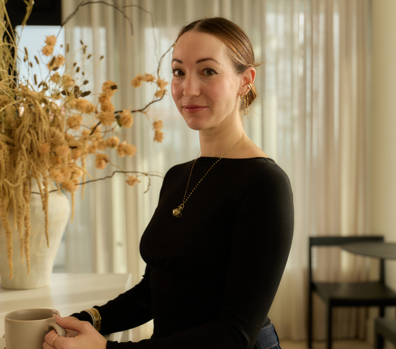 Maxime standing indoors holding a ceramic mug beside a table with a dried floral arrangement.