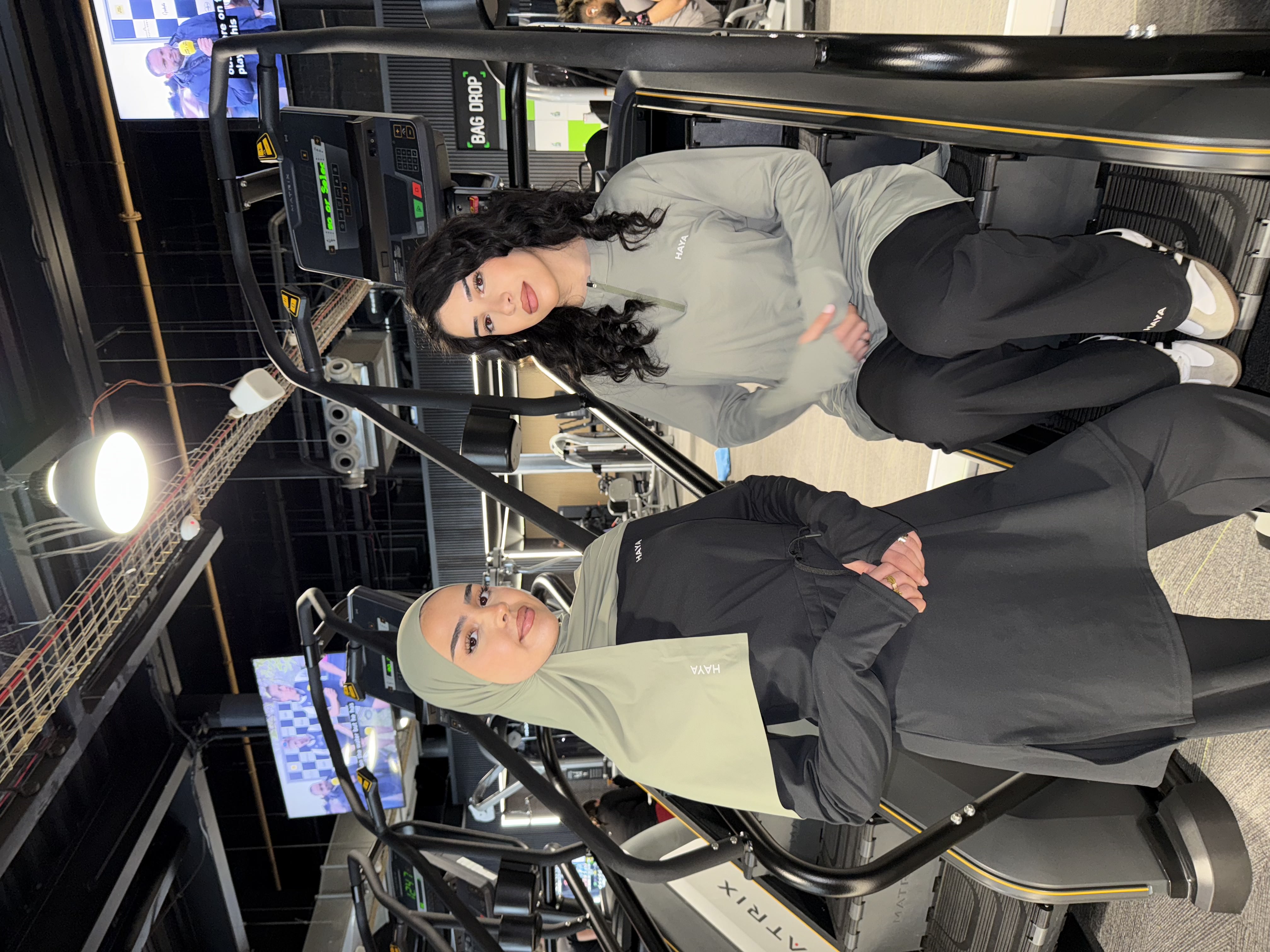 Two women in sportswear posing in a gym with exercise equipment.