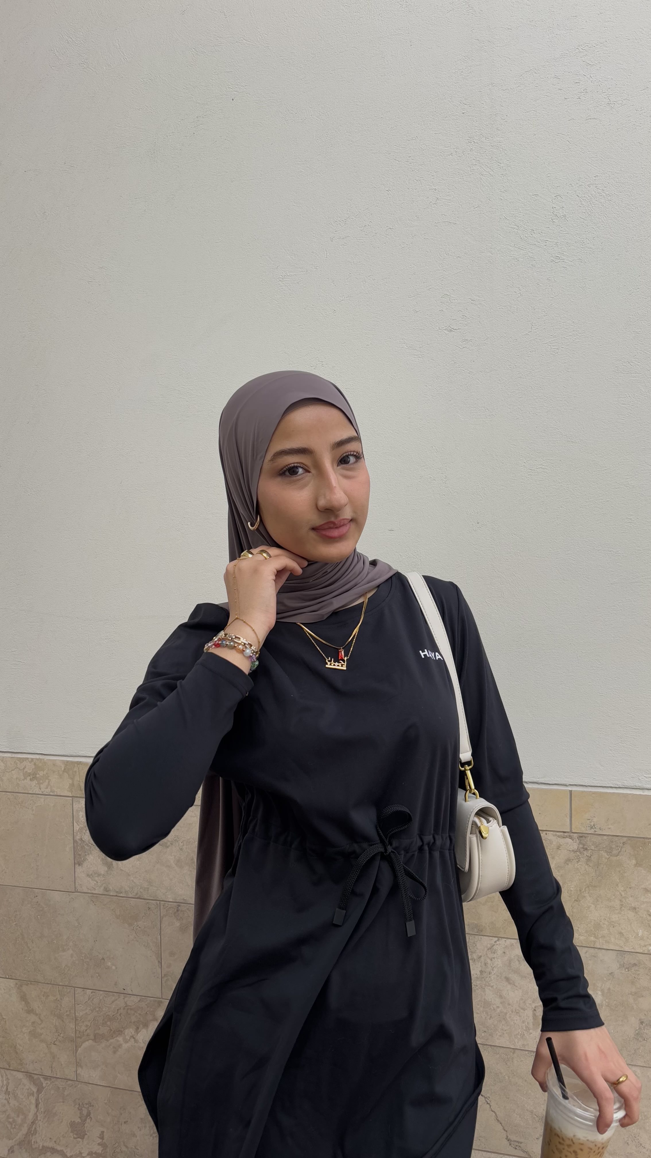 Person in a black outfit and gray headscarf holding a drink.