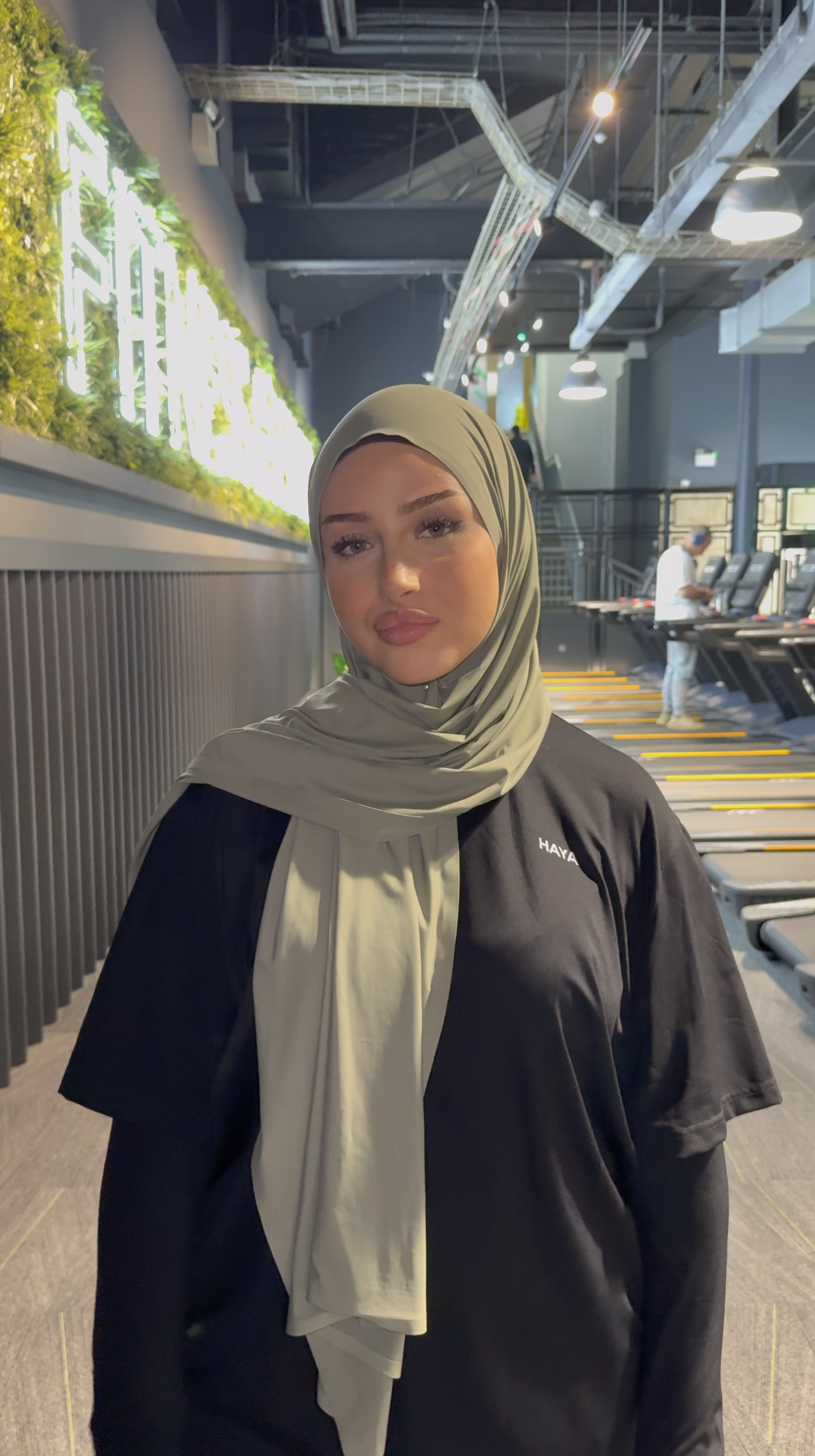 Woman in a gym wearing a hijab and black shirt, standing near treadmills.