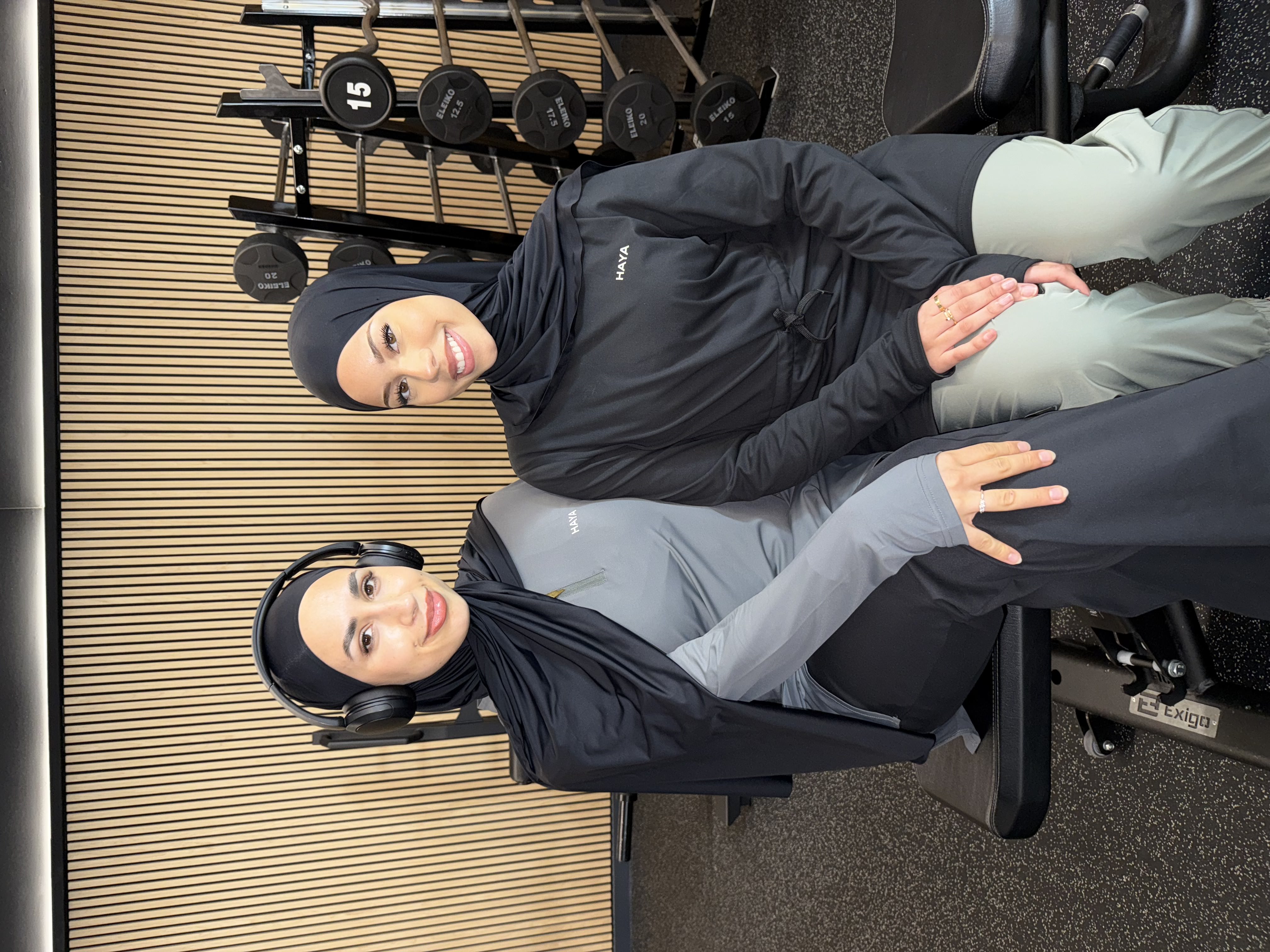 Two women in sportswear sitting on a gym bench, smiling, with weights in the background.