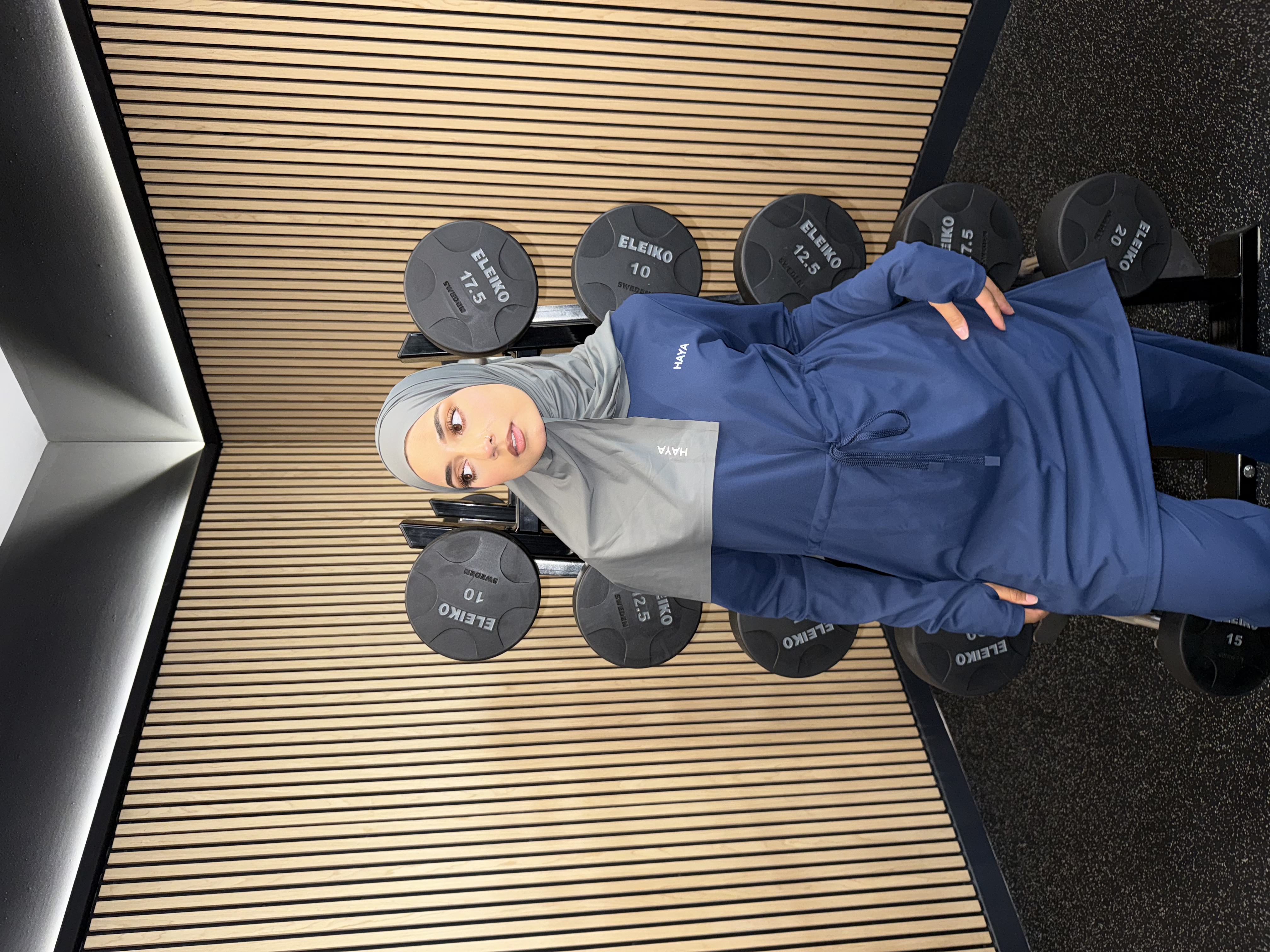 Person in blue sportswear and hijab standing by gym weights.