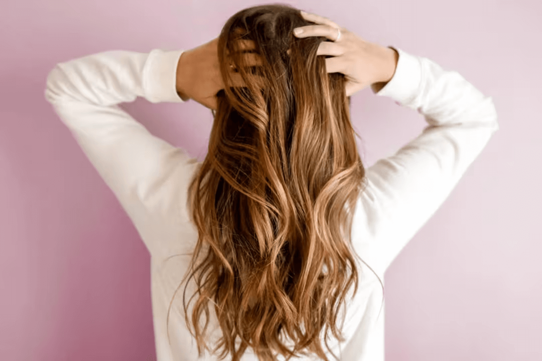 Person with long wavy hair, hands on head, against a pink background.