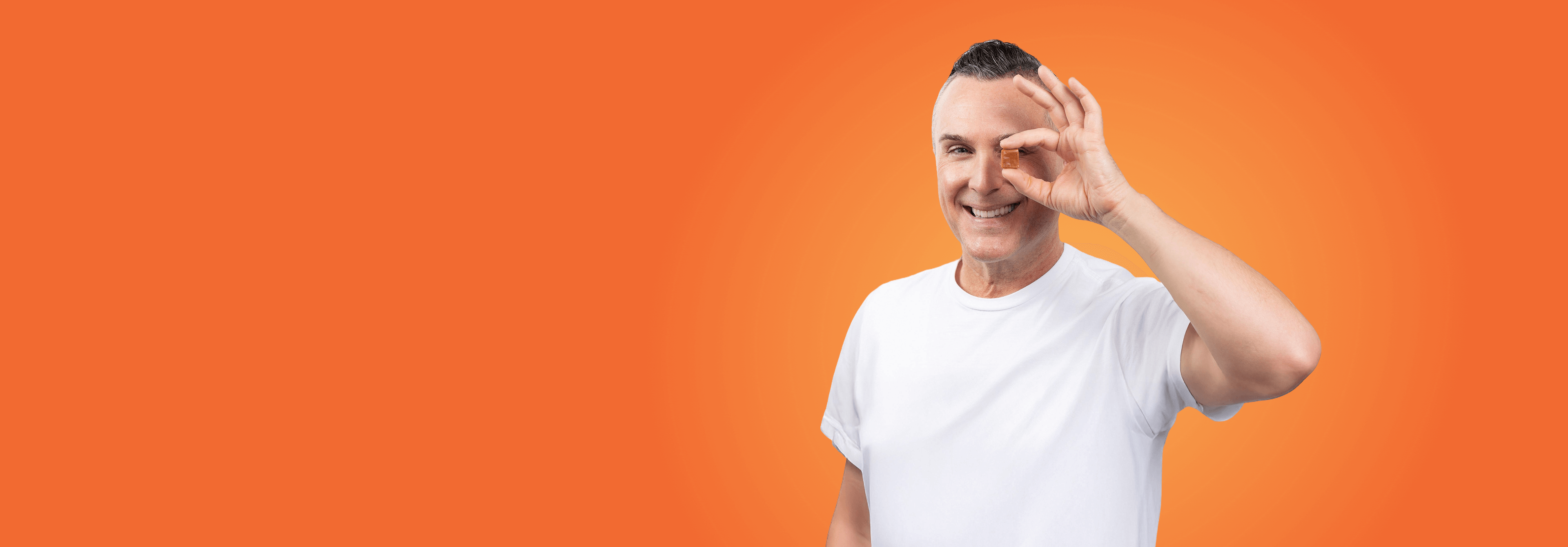 A smiling man in a white t-shirt makes an OK gesture over his eye against an orange background.