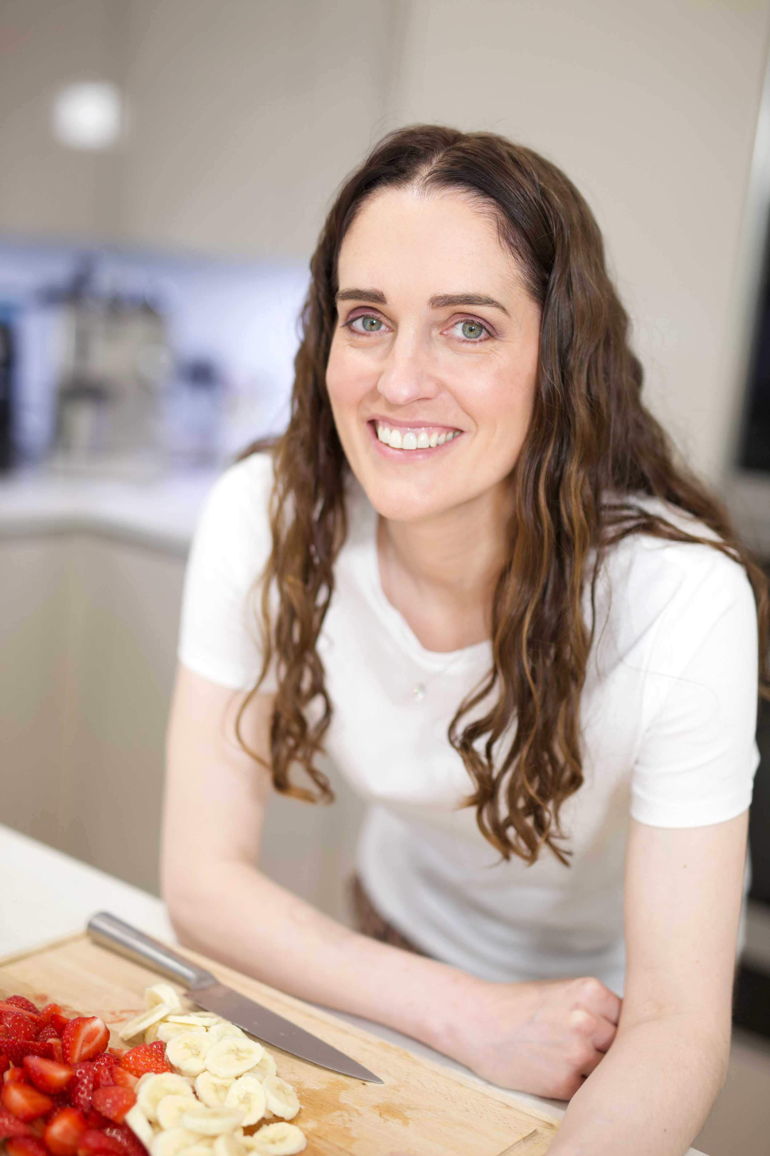 A smiling woman with long brown hair leans over a cutting board with sliced strawberries and bananas.