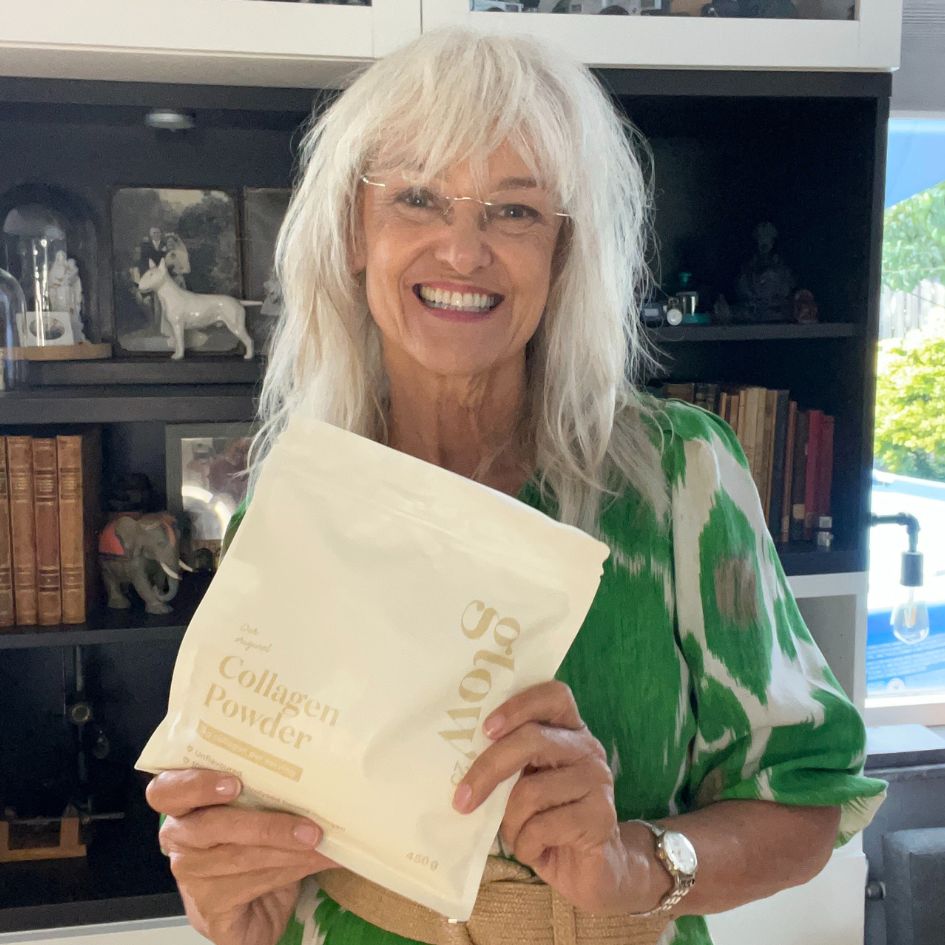 A smiling woman with white hair and glasses holds a white pouch of collagen powder.