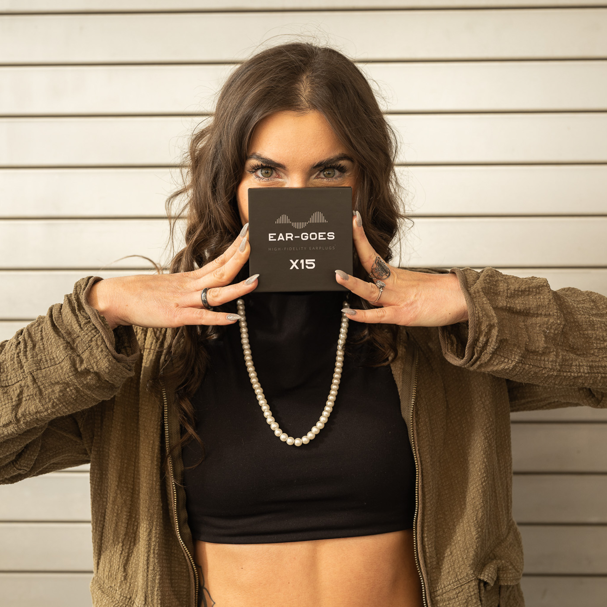 Person holding an 'Ear-Goes X15' box near their face against a slatted wall.