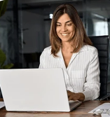 A smiling woman in a white collared shirt sits at a desk using a laptop computer.