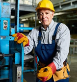 An older male factory worker in a hard hat and gloves stands next to blue machinery.