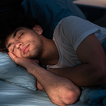 A young man with dark hair sleeps peacefully in bed, his head on a blue pillow.