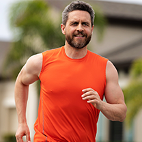 A middle-aged man with a beard wearing an orange sleeveless shirt jogging outdoors.