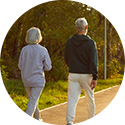 Rear view of an older couple walking together on a paved path through a sunlit, wooded area.
