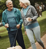 An older man and woman in athletic wear smile at each other while walking a dog in a park.