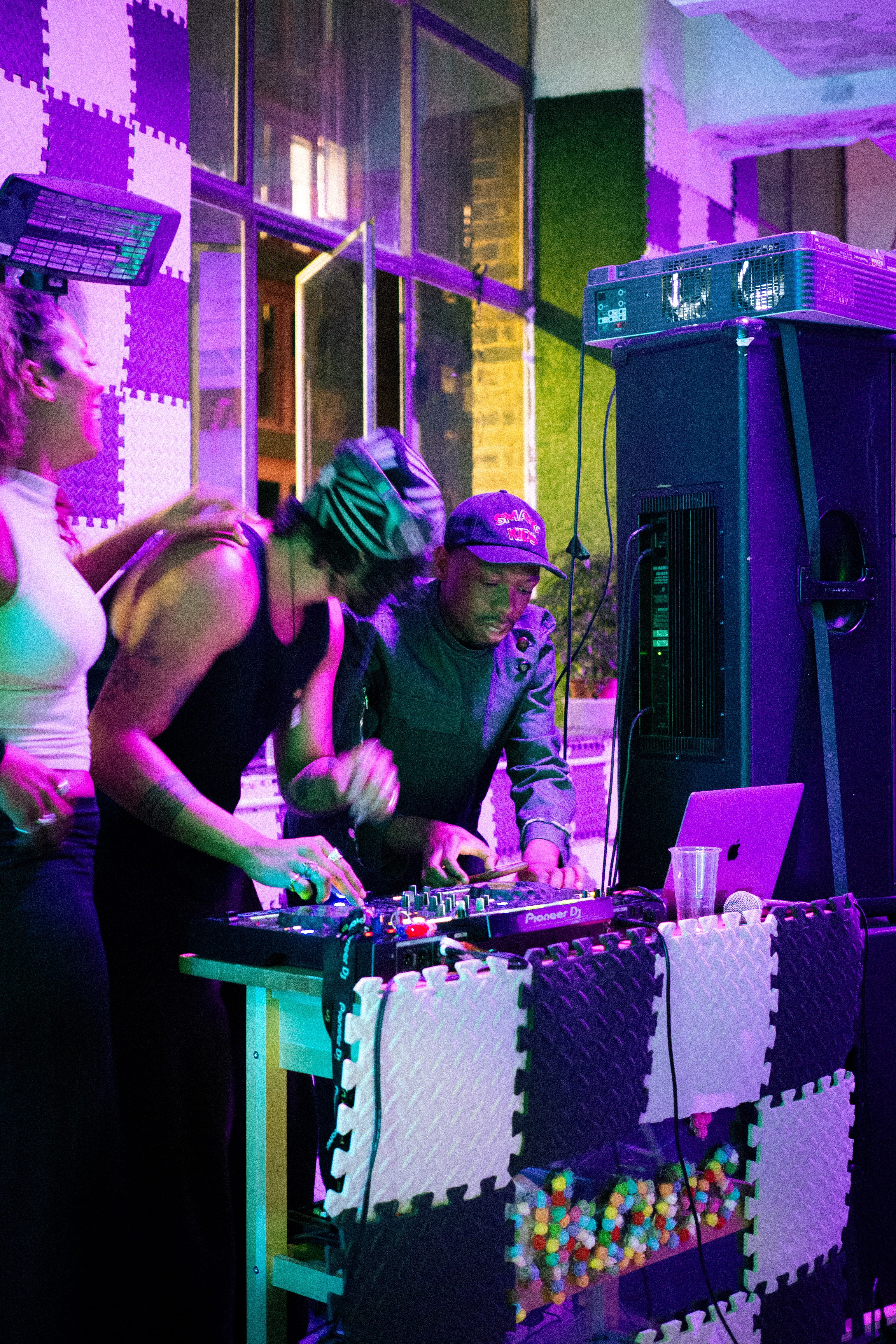 Three people DJing with equipment in a colorful, illuminated room.