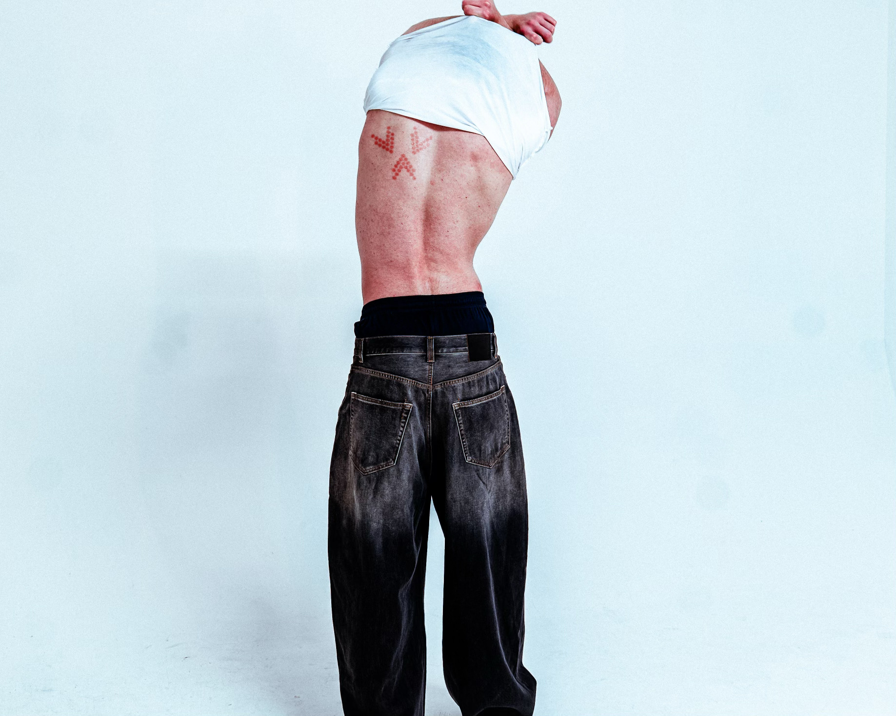 Person removing a white shirt, showing a tattooed back, wearing jeans.