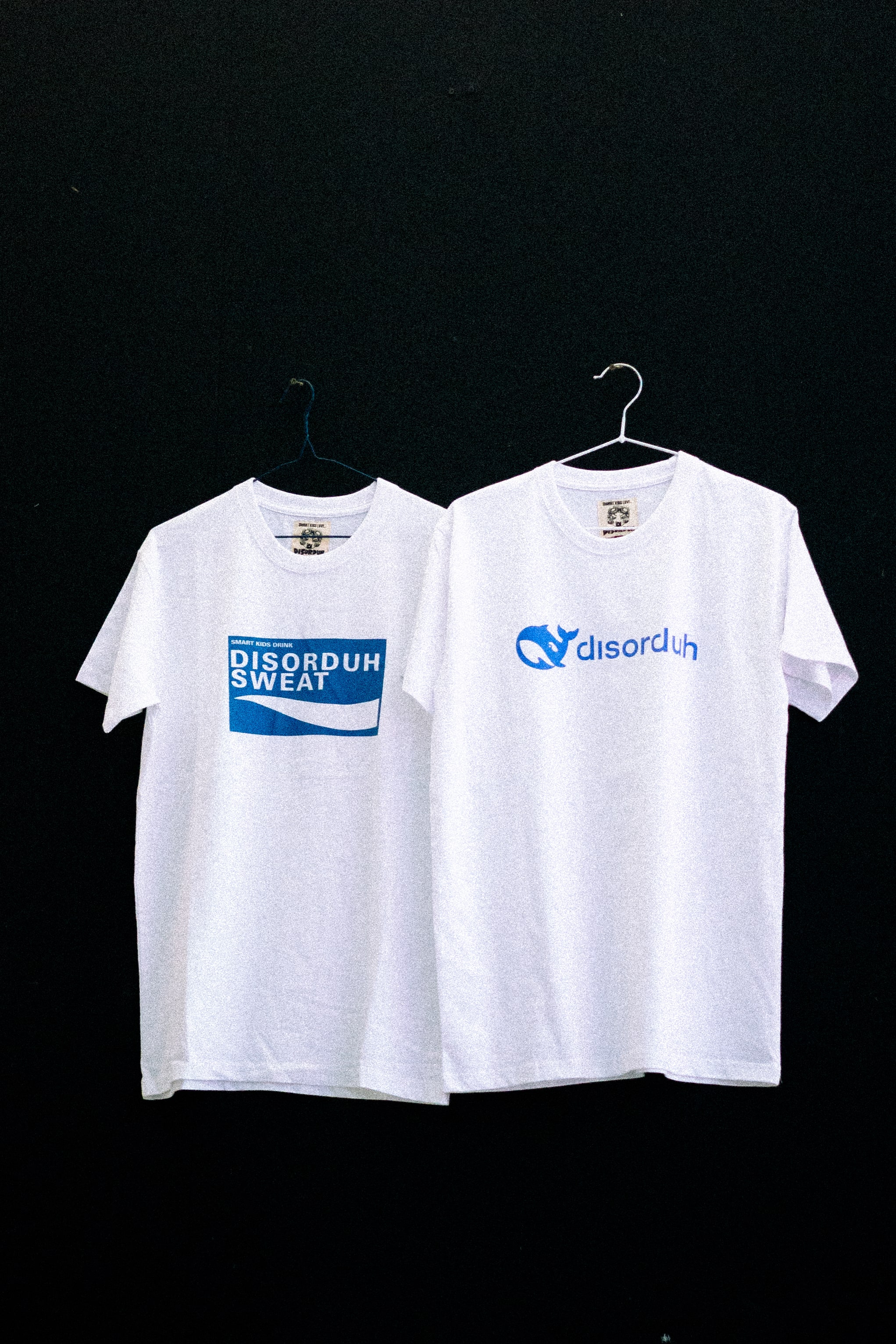 Two white T-shirts with blue logos hanging on black background.