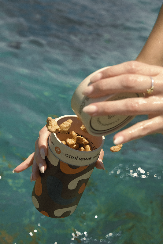 Hands opening a colorful container with snacks near water.