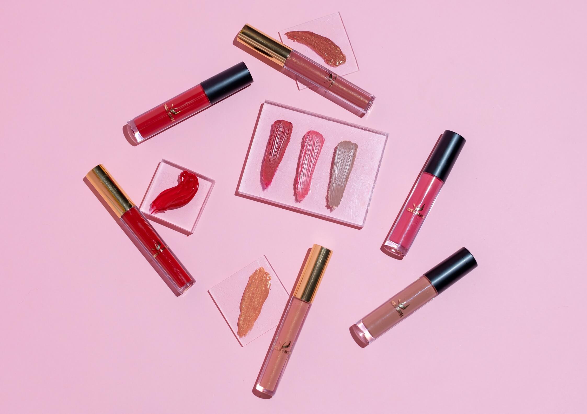 Assorted lip gloss tubes and swatches on a pink background.