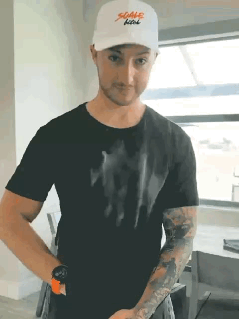 Man in black shirt and cap with arm tattoos, standing indoors.