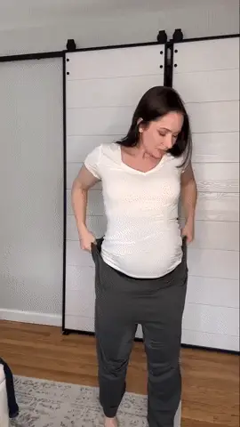 Woman in white shirt adjusting oversized gray pants indoors.
