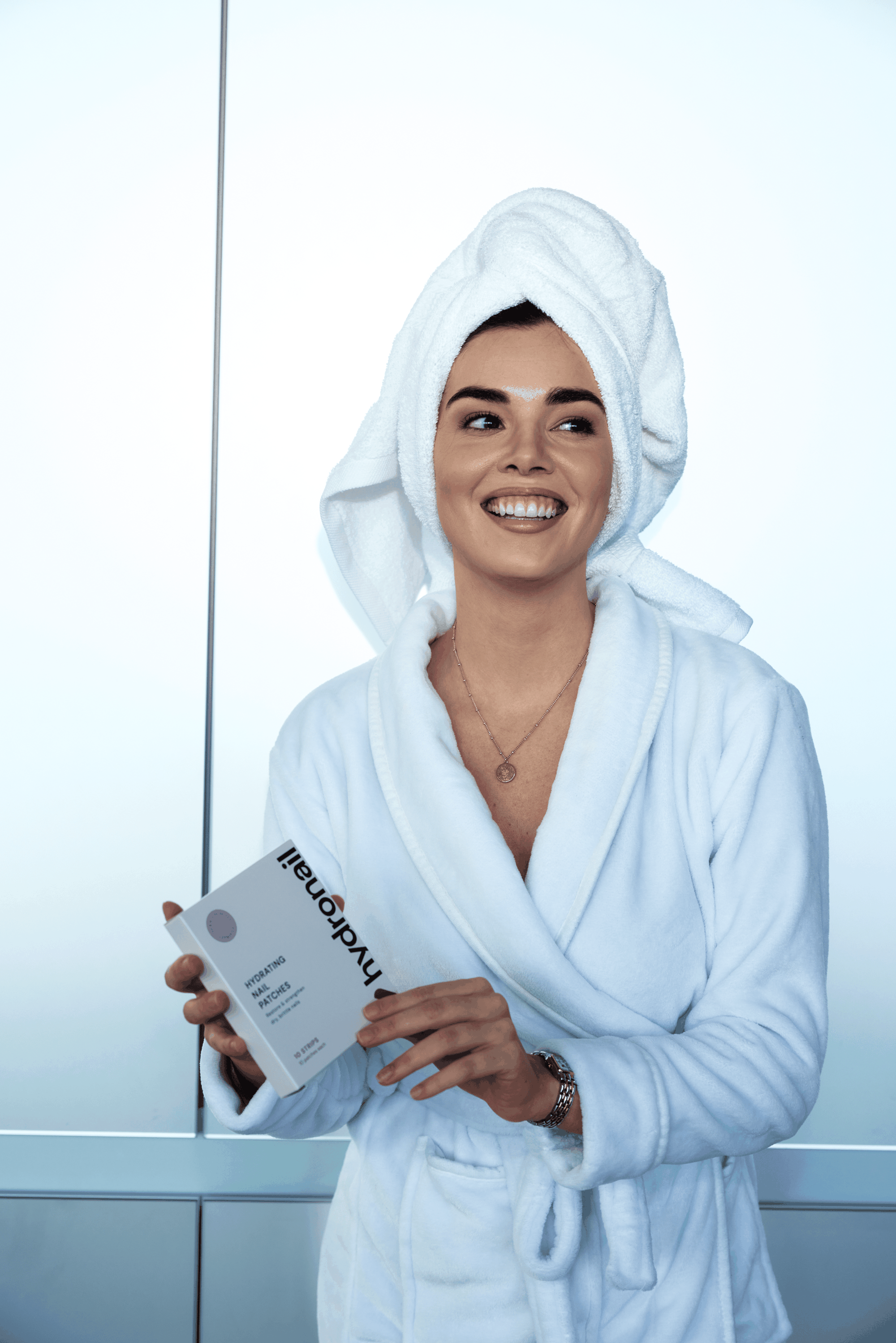 A smiling woman in a bathrobe and hair towel holds a box of Hydronail nail patches.